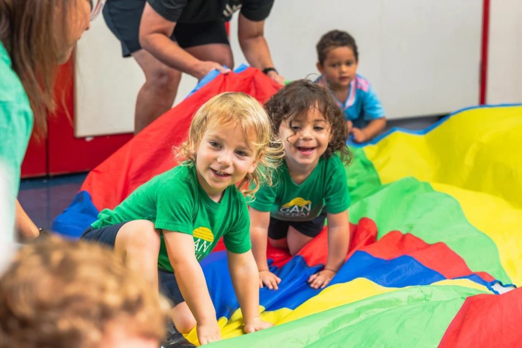 Smiling Gan Sinai preschoolers crawling across a colorful play parachute